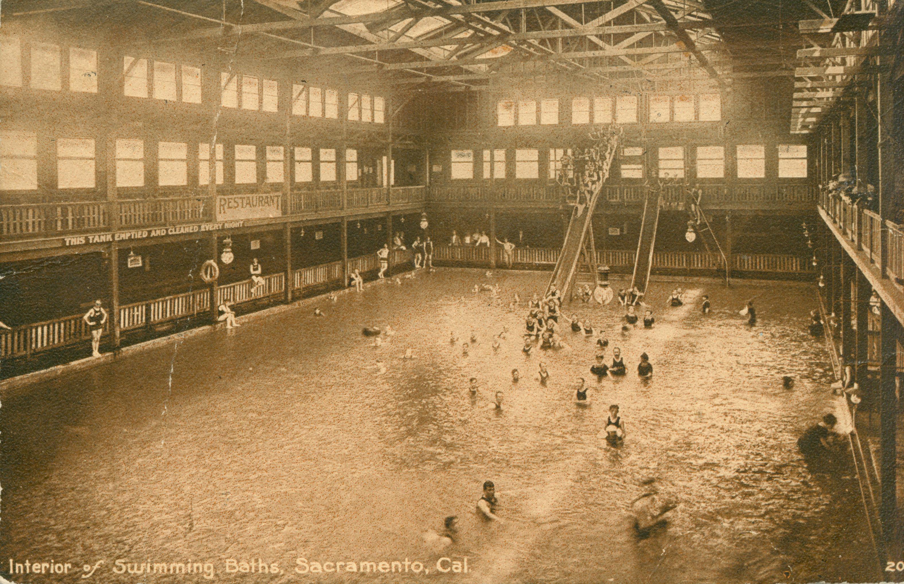 This postcard shows the interior of an indoor swimming pool, with several people in the water. There are two water slides at the far end of the pool.